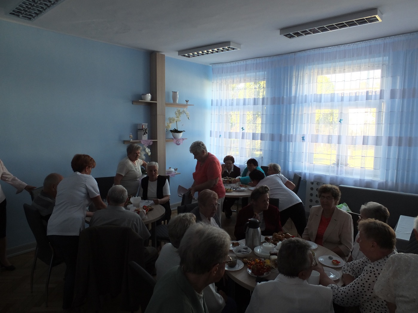 Uczestnicy Dziennego Domu Pomocy podczas poczęstunku na otwarcie Domu, uczestnicy siedzą przy stolikach w grupach, rozmawiają, jedzą ciasto