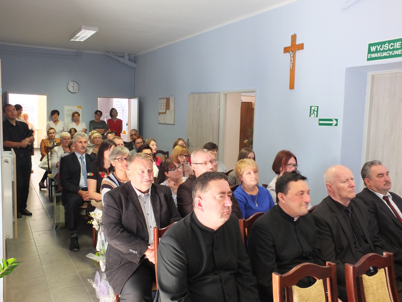 Grupa gości podczas otwarcia Dziennego Domu Pomocy w pomieszczeniu Domu, goście siedzą na krzesłach