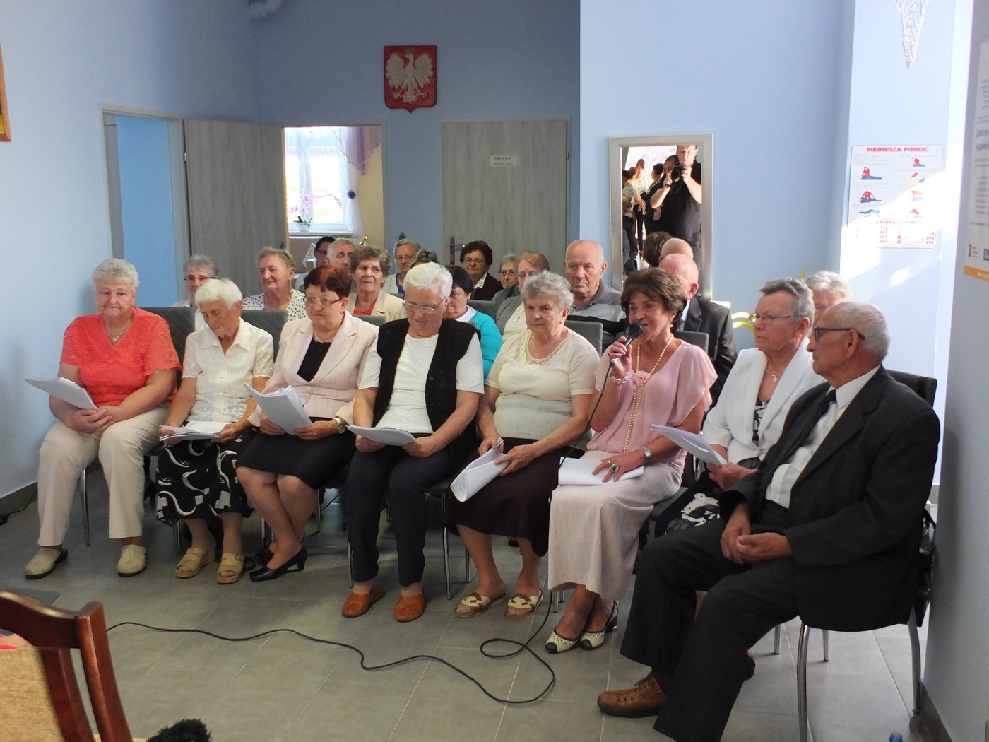 Grupa uczestników Dziennego Domu Pomocy podczas otwarcia Domu - część artystyczna, osoby siedzą na krzesłach, recytują wiersze