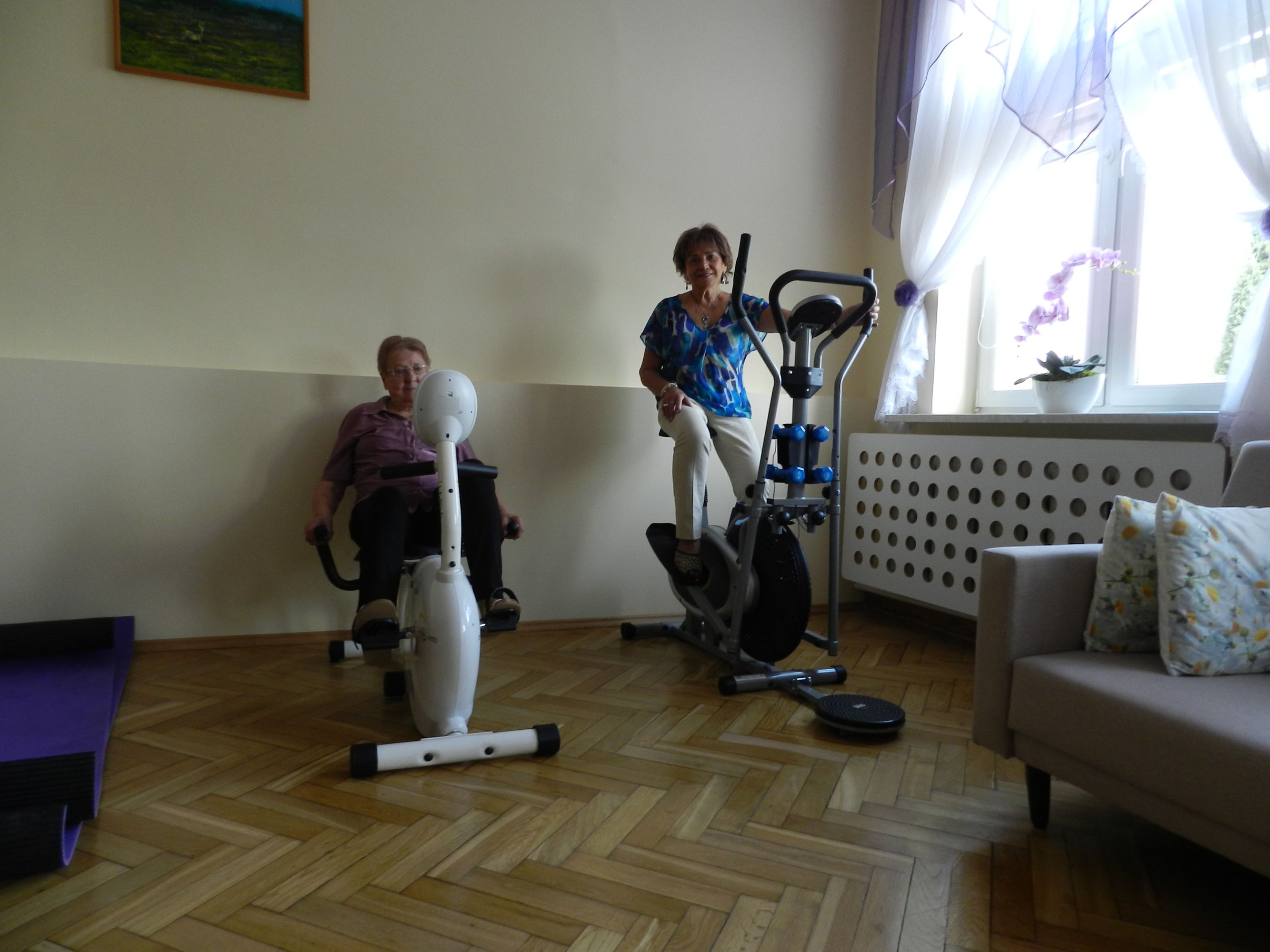 2 uczestniczki Dziennego Domu Pomocy podczas ćwiczeń na rowerku stacjonarnym i orbitreku, w tle okno