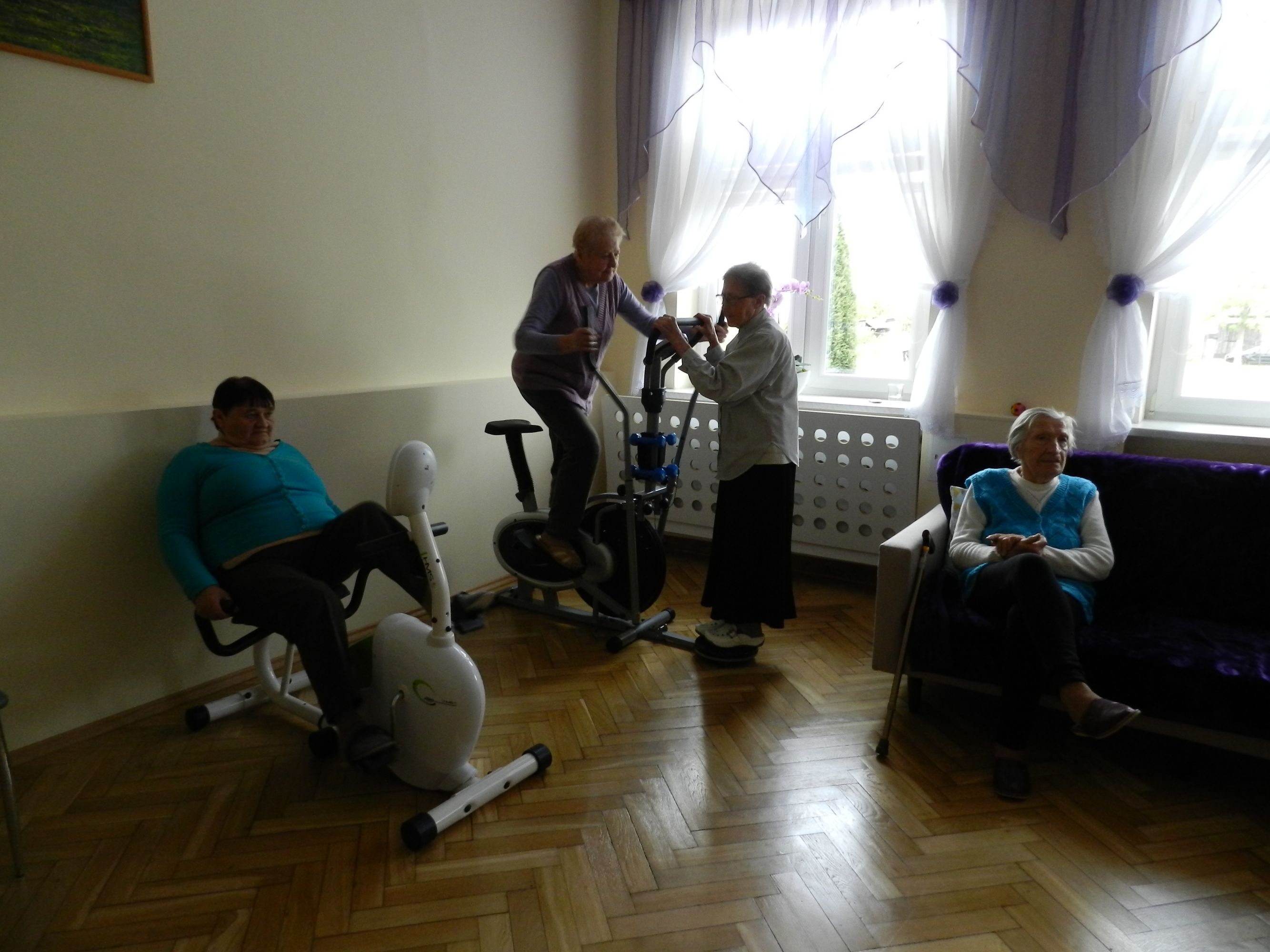 4 uczestników Dziennego Domu Pomocy w sali do rehabilitacji, 1 osoba ćwiczy na rowerku stacjonarnym, 1 osoba na orbitreku, 1 osoba stoi przy orbitreku, 1 osoba odpoczywa na kanapie mieszczącej się w pokoju, w tle okna