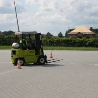 Uczestnik kursu poruszający się wózkiem widłowym po placu manewrowym, ubrany w kask i kamizelkę ochronną, w tle widać krzewy oraz dach budynku