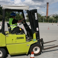 Uczestnik kursu poruszający się wózkiem widłowym w trakcie wykonywania manewru na placu, ubrany w kask i kamizelkę ochronną, w tle widać, pachołki oraz pasy namalowane na placu