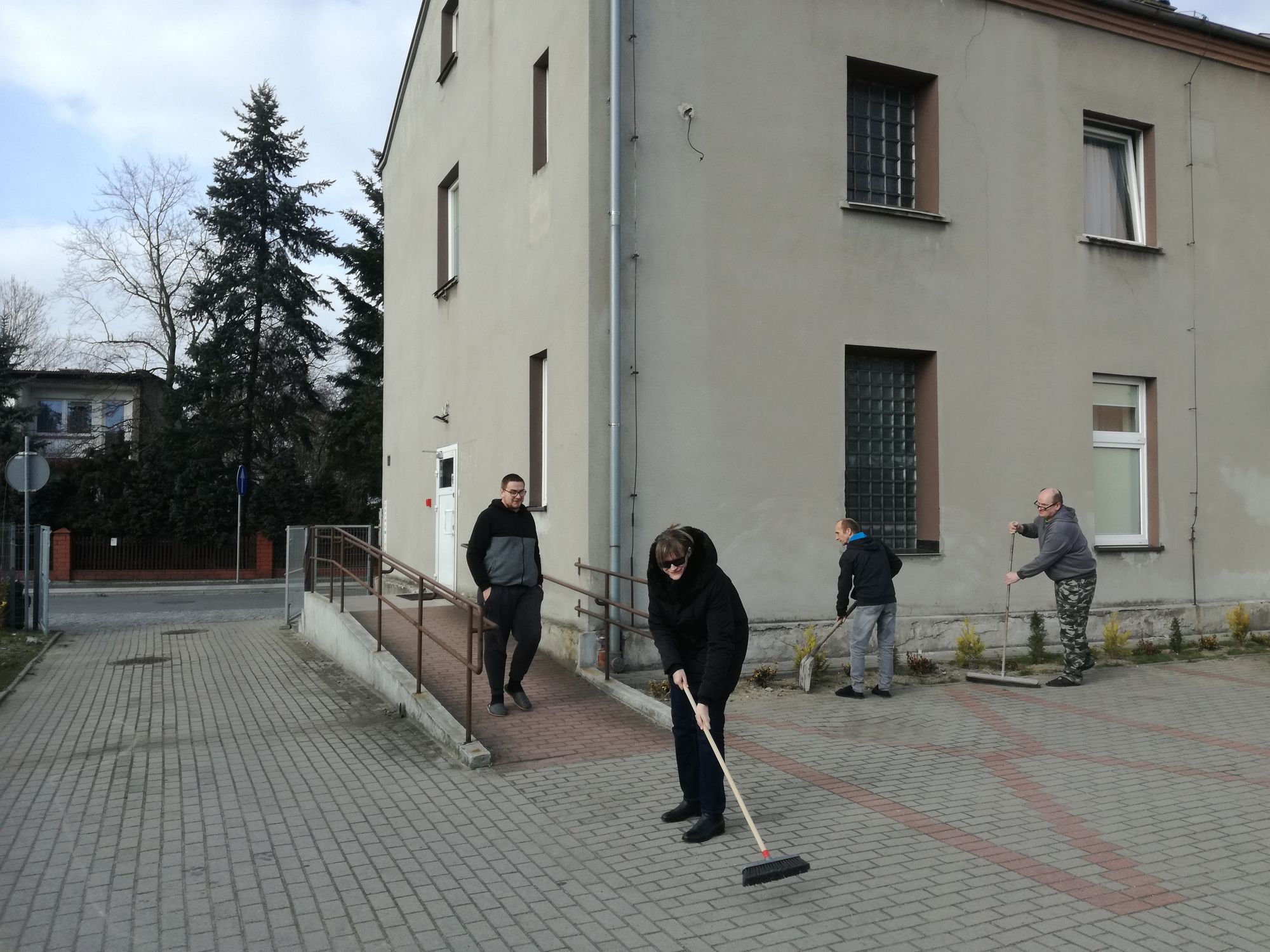 Trzej mieszkańcy mieszkań wspomaganych podczas prac porządkowych wokół budynku (zamiatają), jeden mieszkaniec schodzi z podjzdu do wózków inwalidzkich